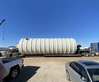Large underground fiberglass water storage tank secured on flatbed trailer for transport to construction site, with crane and trucks in background